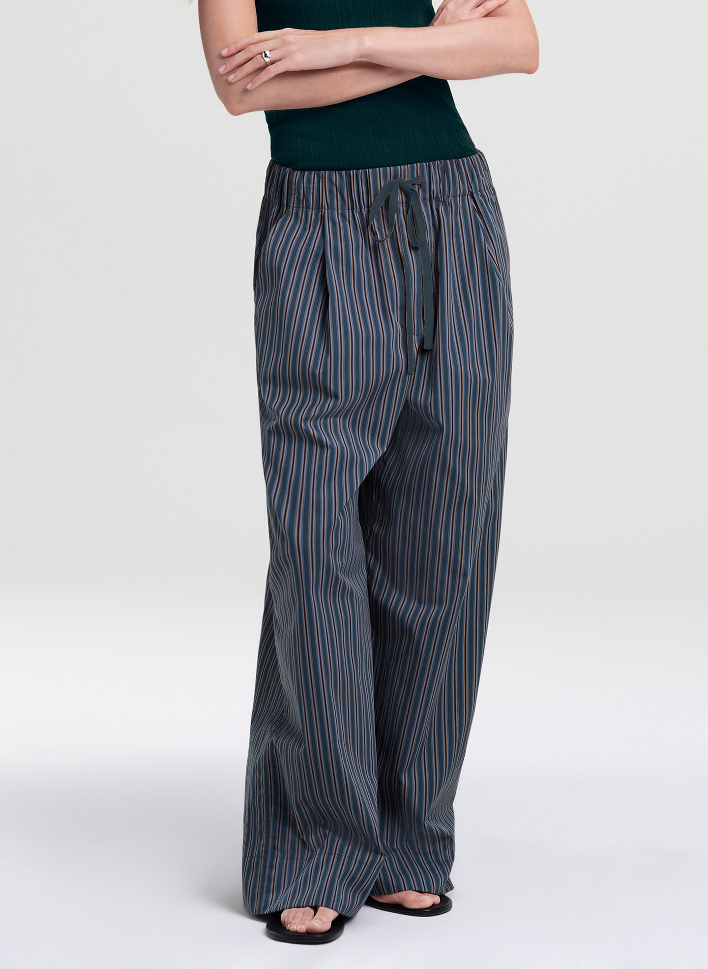 A model wearing wide-leg drawstring pants with vertical blue and orange pinstripes on a dark gray base, paired with a dark green top and black sandals, against a light gray studio background.