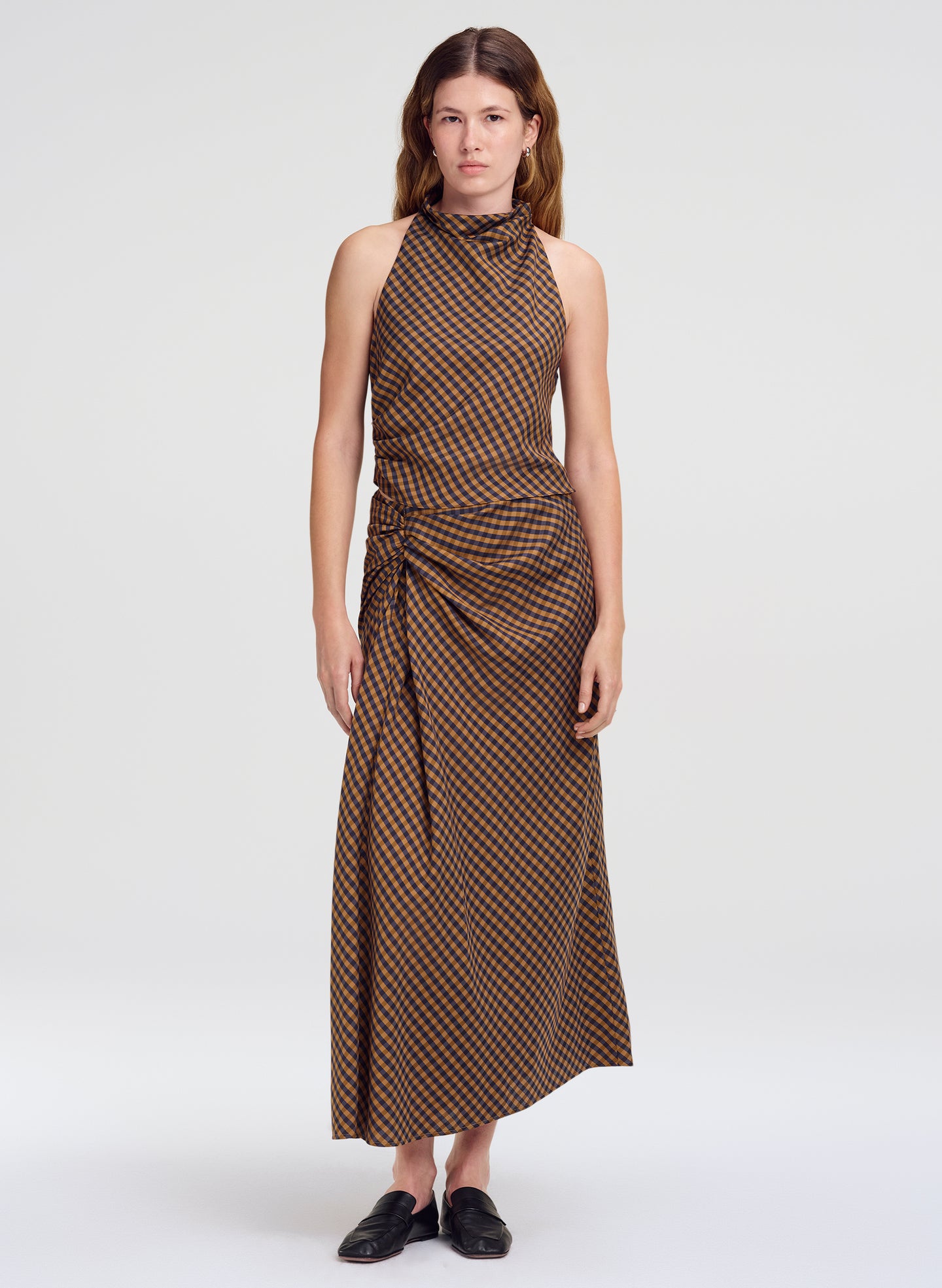 A woman modeling a sleeveless top and skirt with a brown and beige geometric pattern, standing against a light gray studio background.