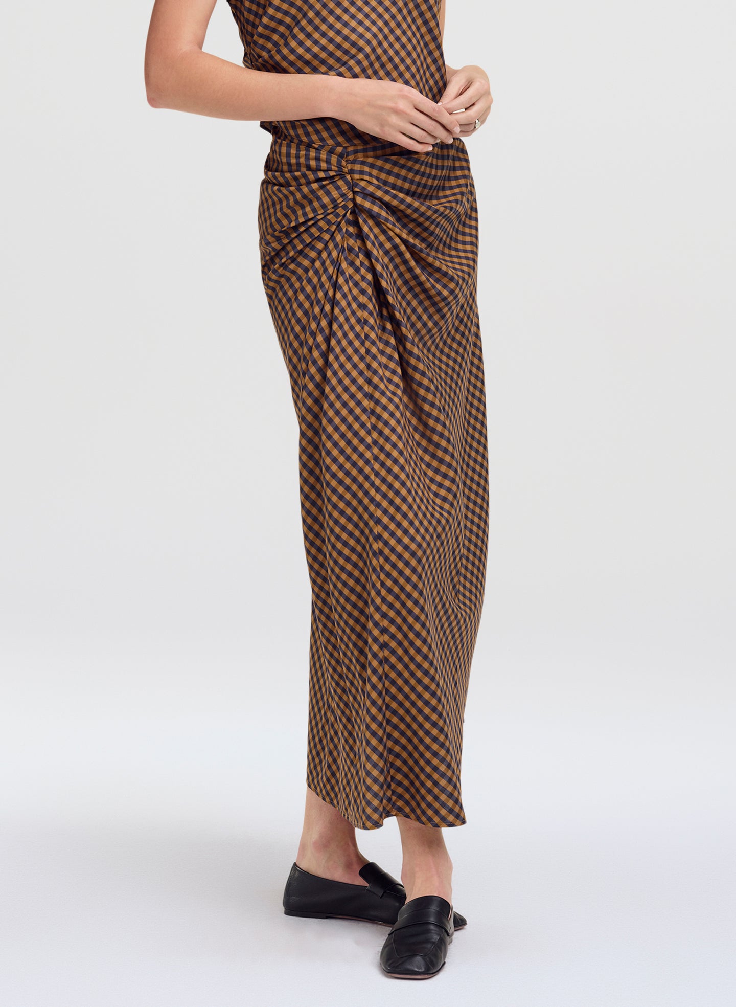 A model wearing a brown and black checkered wrap skirt with a gathered waist, paired with black flat shoes, against a light gray studio background.