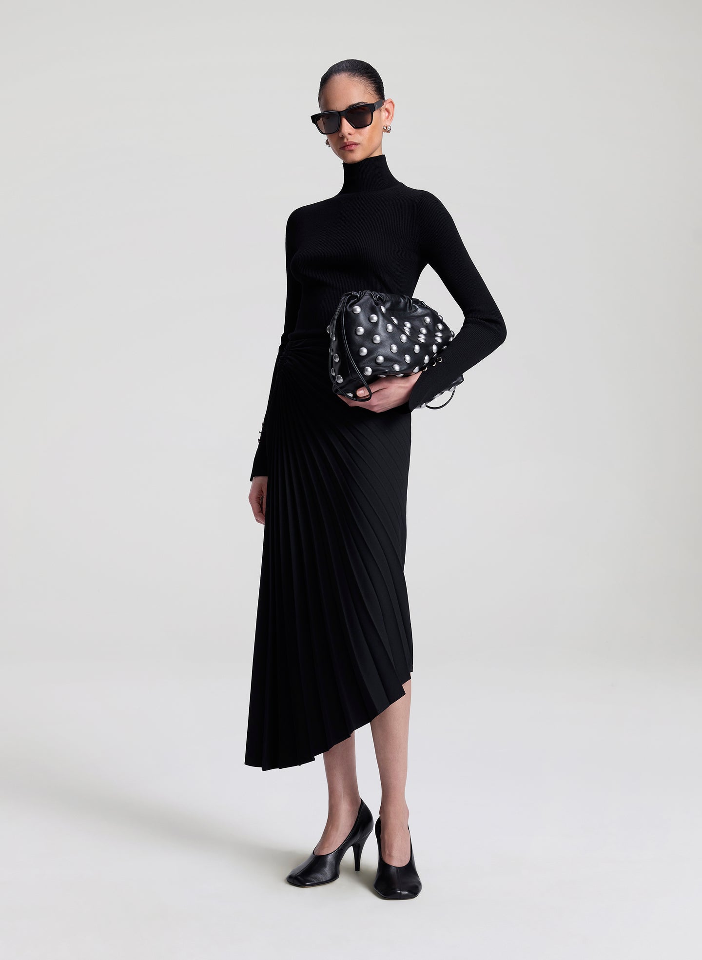 Woman in black turtleneck and pleated skirt holding a polka dot handbag on a white background