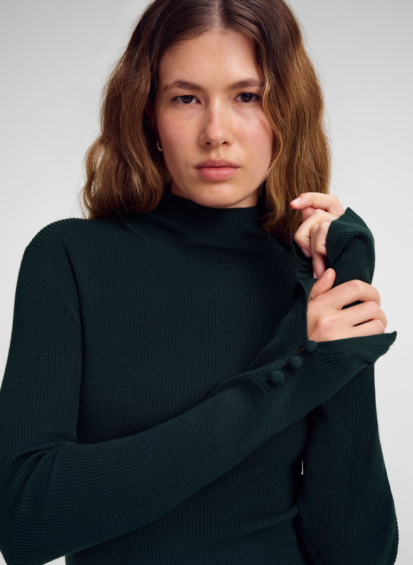 A woman wearing a dark forest green ribbed mock neck sweater with long sleeves and decorative buttons on the cuffs, posed against a neutral light gray background.