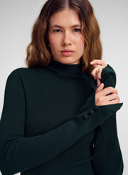 A woman wearing a dark forest green ribbed mock neck sweater with long sleeves and decorative buttons on the cuffs, posed against a neutral light gray background.