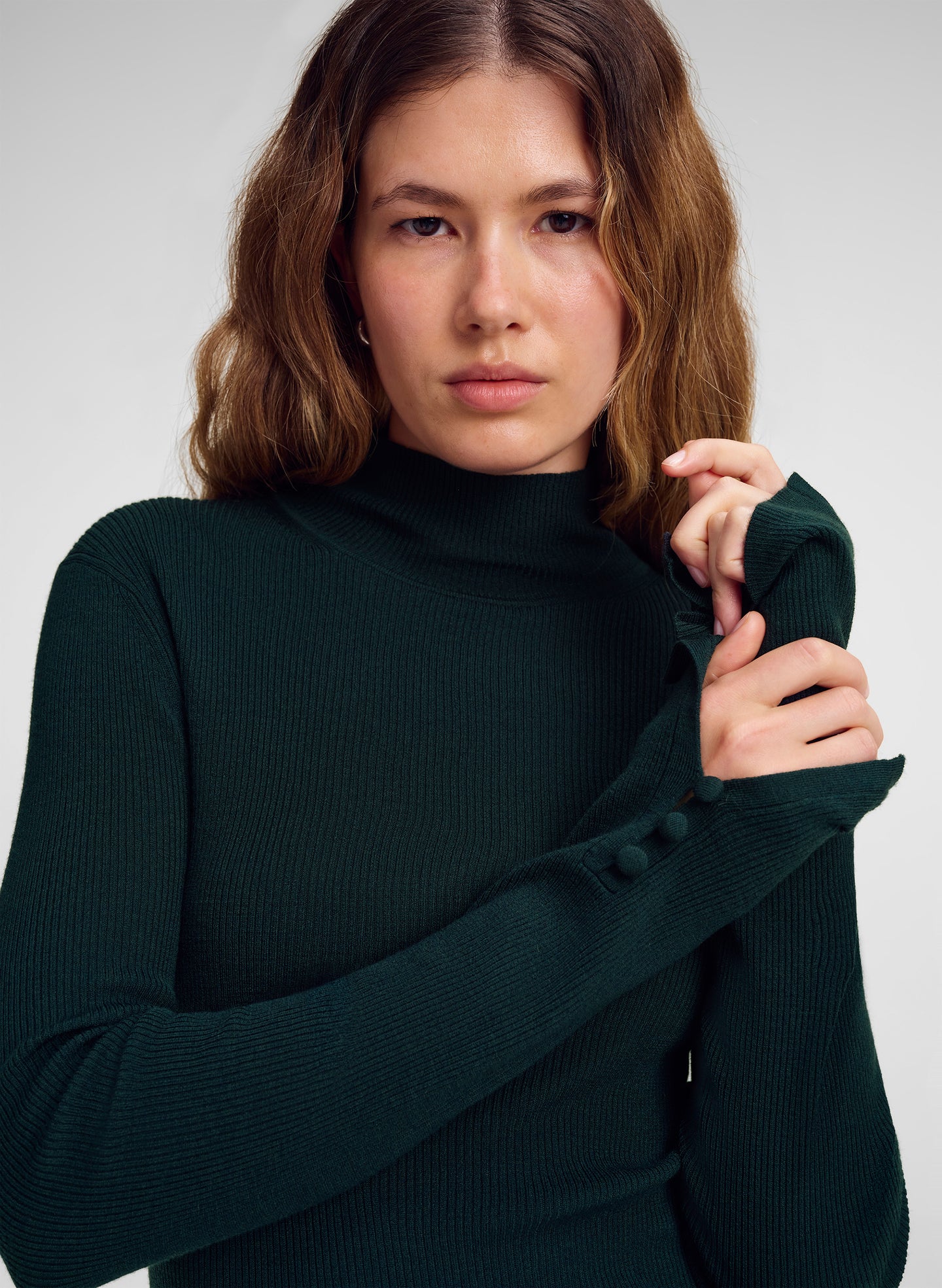 A woman wearing a dark forest green ribbed mock neck sweater with long sleeves featuring decorative buttons on the cuffs, posed against a neutral light gray background.