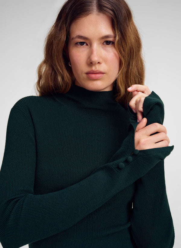 A woman wearing a dark forest green ribbed mock neck sweater with long sleeves featuring decorative buttons on the cuffs, posed against a neutral light gray background.