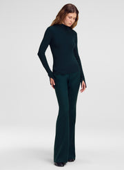 A woman modeling a dark forest green ribbed knit two-piece set consisting of a mock-neck long-sleeve top and matching wide-leg trousers, shot against a light gray studio background.