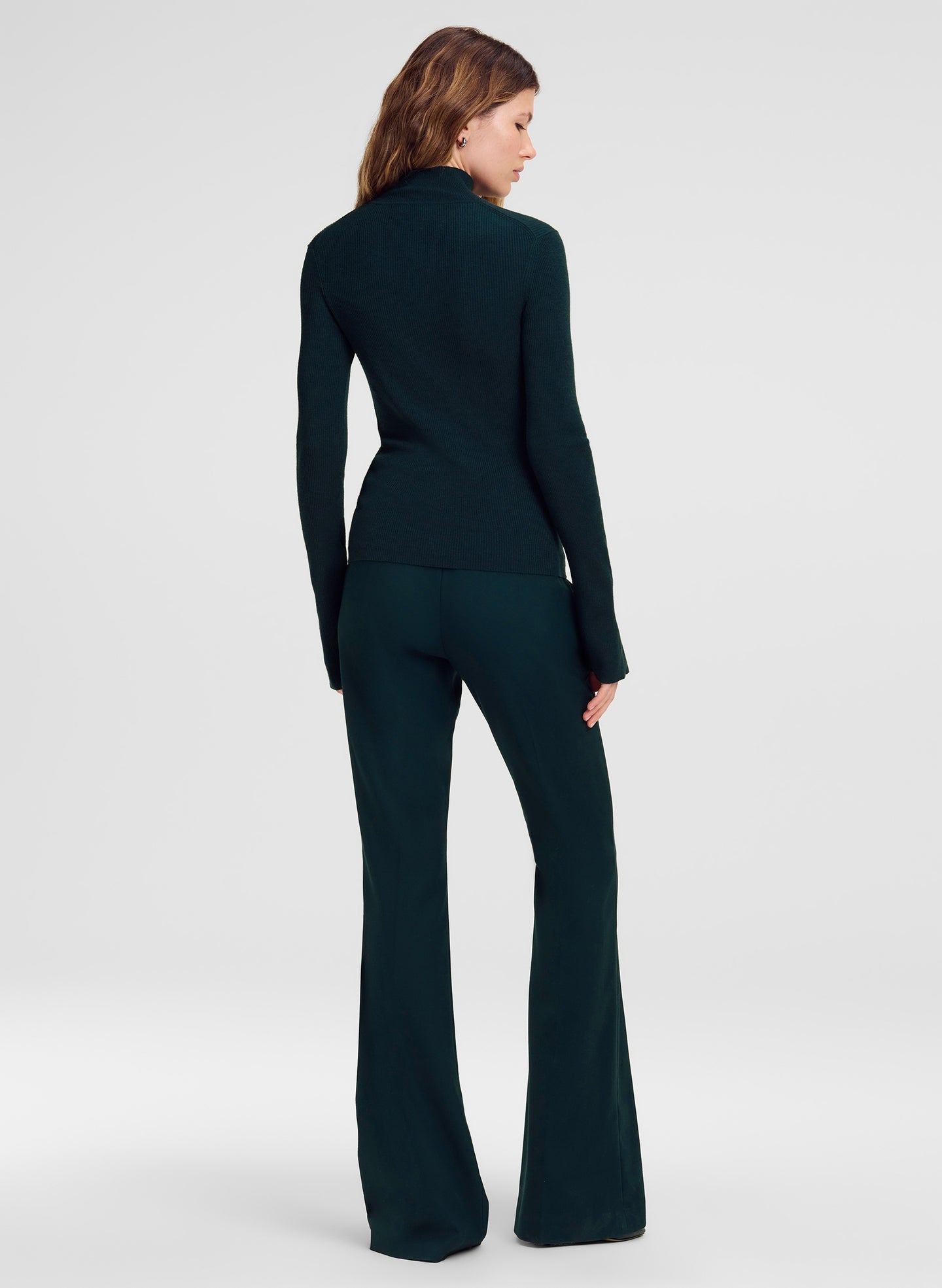 A woman shown from the back wearing a dark teal-green long-sleeve mock-neck top and matching flared pants against a light gray studio background.