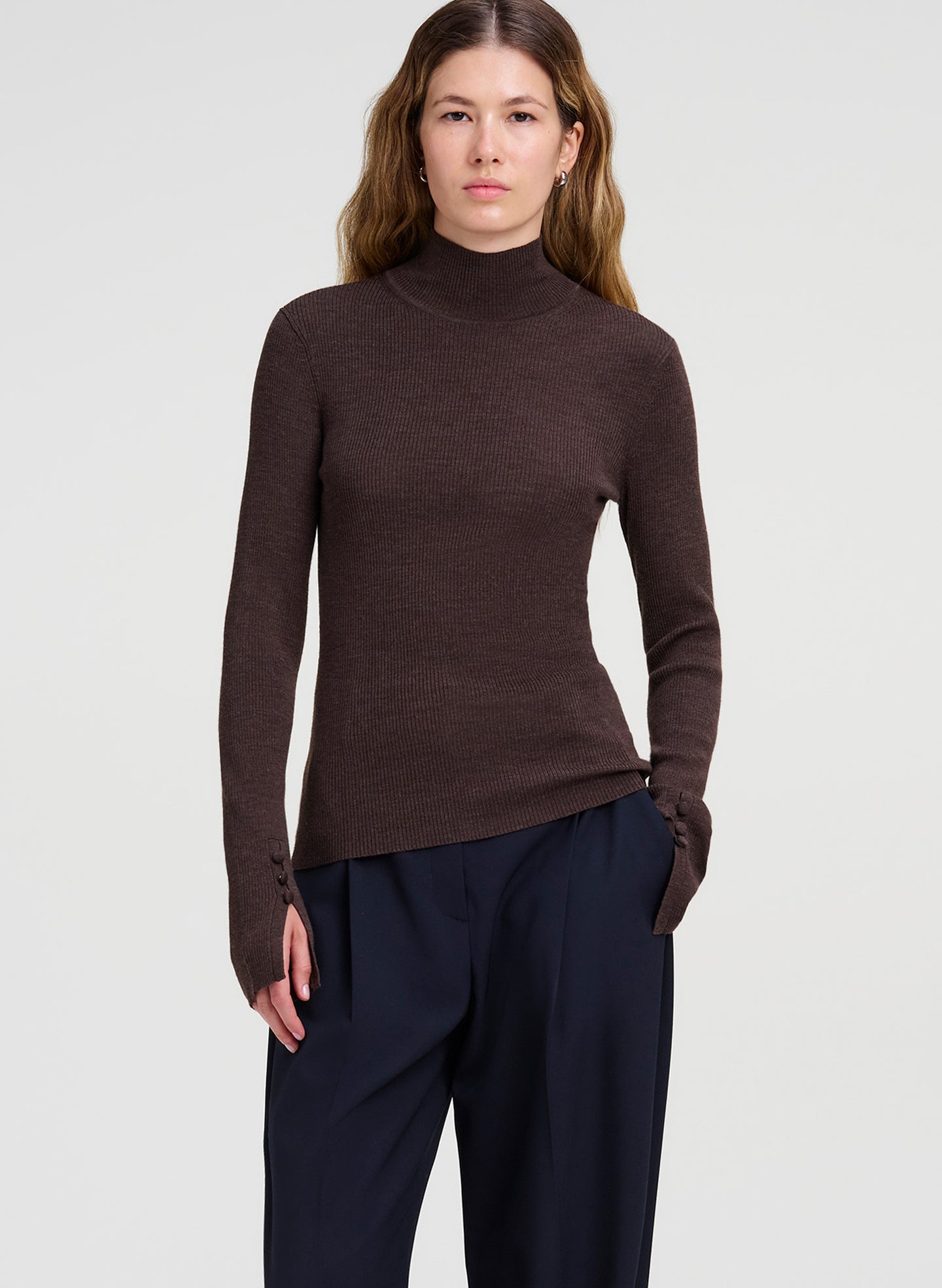 A woman modeling a dark brown ribbed turtleneck sweater with long sleeves and dark navy trousers, photographed against a light gray studio background.