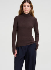 A woman modeling a dark brown ribbed turtleneck sweater with long sleeves and dark navy trousers, photographed against a light gray studio background.
