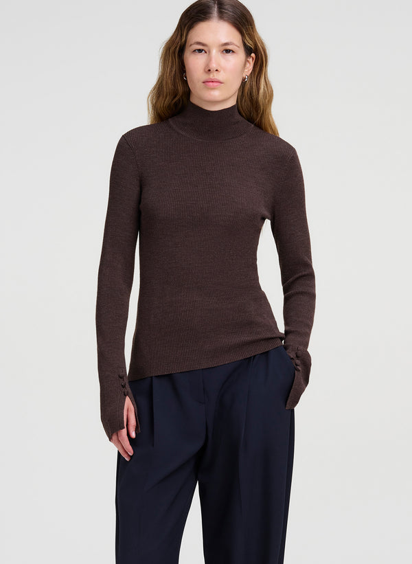 A woman modeling a dark brown ribbed turtleneck sweater with long sleeves and dark navy trousers, photographed against a light gray studio background.