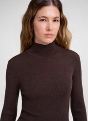 A woman wearing a dark brown ribbed mock neck sweater, shown from the chest up against a light gray studio background.