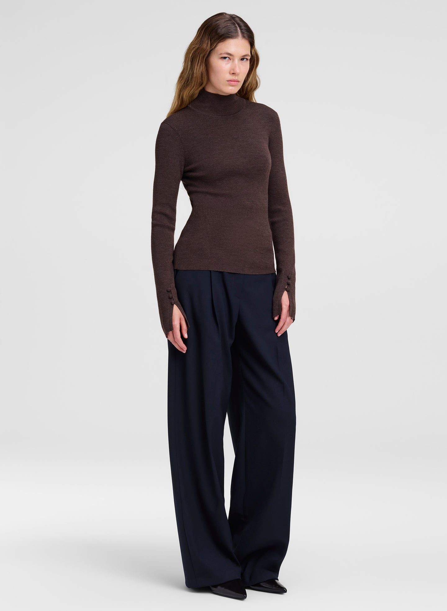 A woman modeling a dark brown mock neck knit sweater paired with dark navy wide-leg trousers, standing against a light gray studio background.