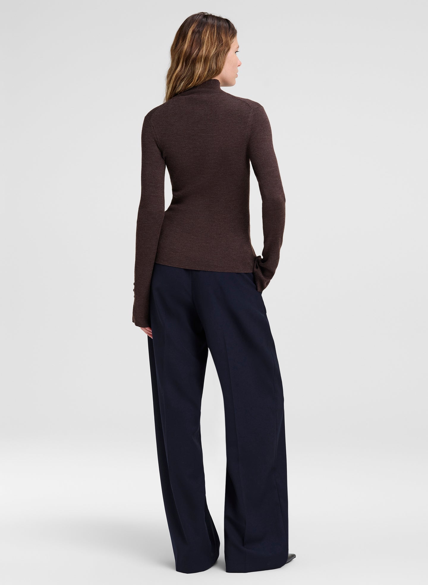 A woman shown from the back wearing a fitted dark brown mock-neck knit top and dark navy wide-leg trousers, photographed against a light gray studio background.