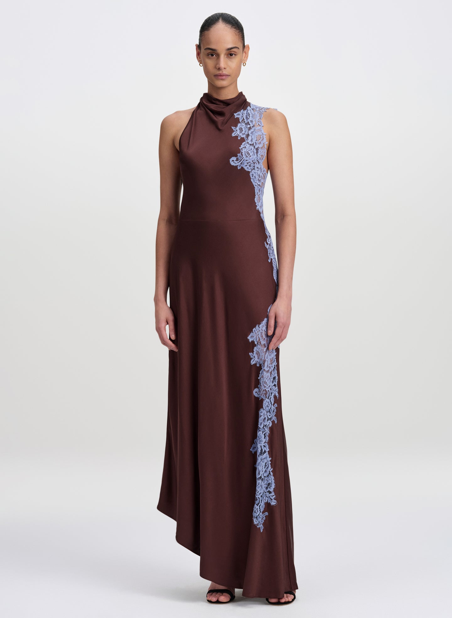 A model wearing a sleeveless, high-neck floor-length gown in deep brown with light blue floral lace applique running down the side, set against a plain light gray studio background.