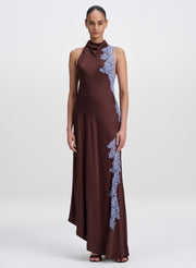 A model wearing a sleeveless, high-neck floor-length gown in deep brown with light blue floral lace applique running down the side, set against a plain light gray studio background.
