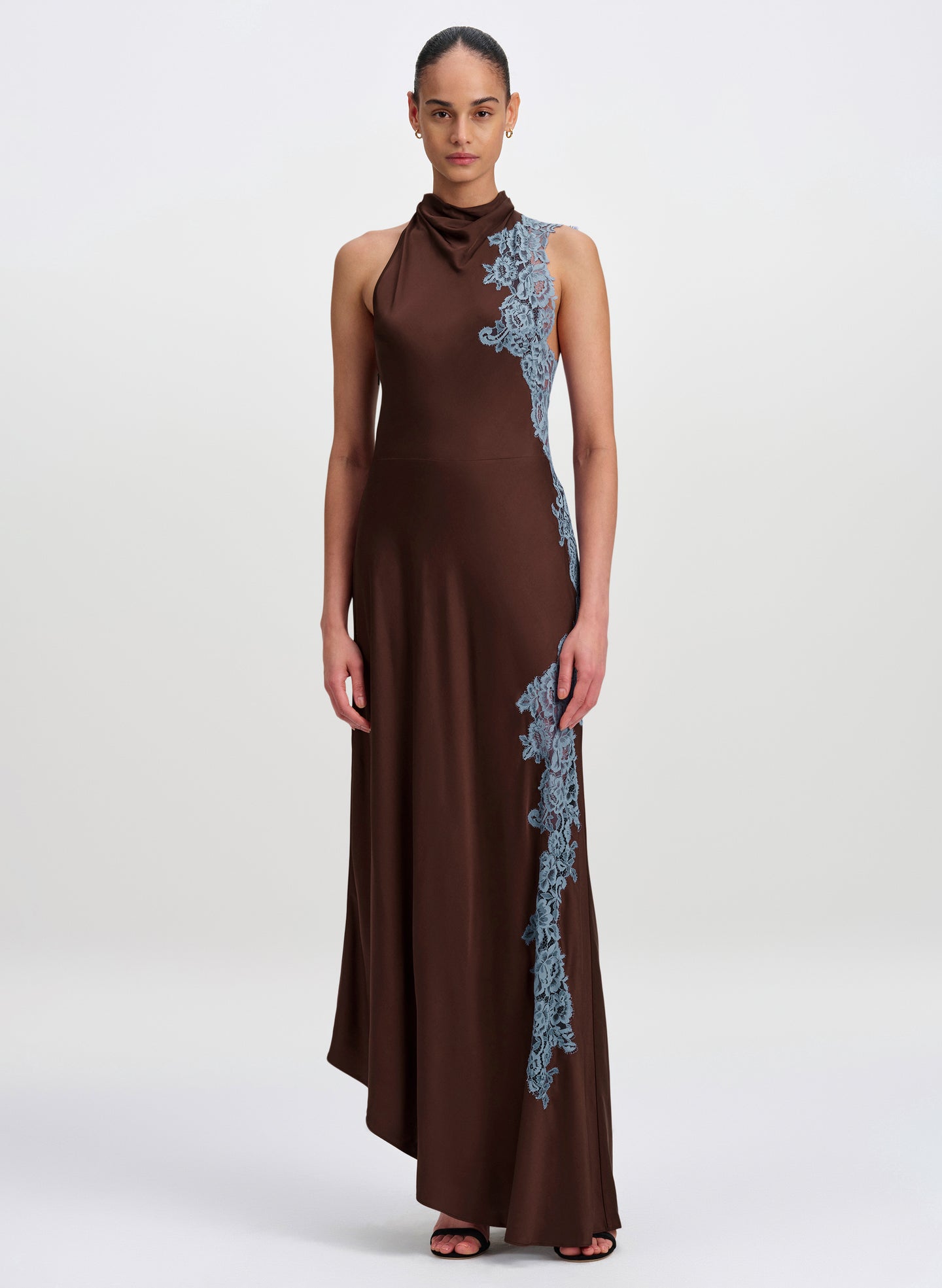 A model wearing a sleeveless high-neck brown maxi dress with light blue floral lace applique down the side, photographed against a neutral light gray studio background.