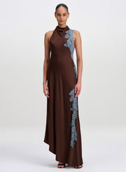 A model wearing a sleeveless high-neck brown maxi dress with light blue floral lace applique down the side, photographed against a neutral light gray studio background.