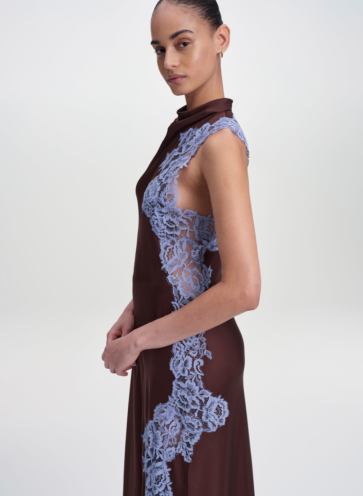 A model wearing a sleeveless high-neck dress in dark brown with light blue floral lace panels, shown from a side profile against a light gray studio background.