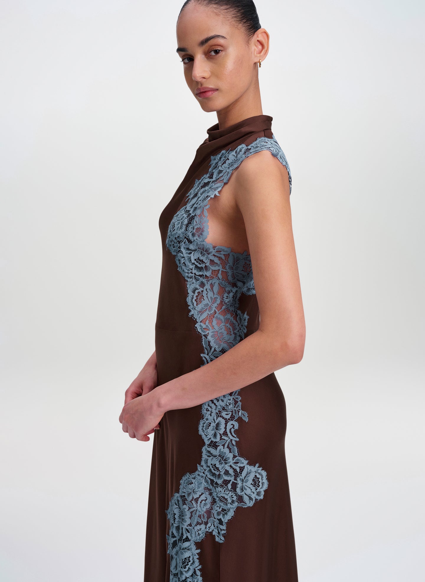 A model wearing a sleeveless high-neck brown dress with prominent dusty blue lace panels on the bodice and side, photographed against a plain light gray studio background.