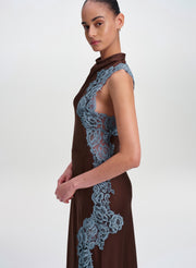 A model wearing a sleeveless high-neck brown dress with prominent dusty blue lace panels on the bodice and side, photographed against a plain light gray studio background.
