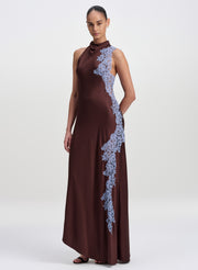 A woman modeling a sleeveless, high-neck floor-length gown in deep brown with a diagonal light blue lace applique detail, photographed against a plain light gray studio background.