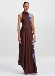 A model wearing a sleeveless, high-neck, floor-length brown dress with light blue floral lace applique detailing along the shoulder and side, set against a plain light gray studio background.