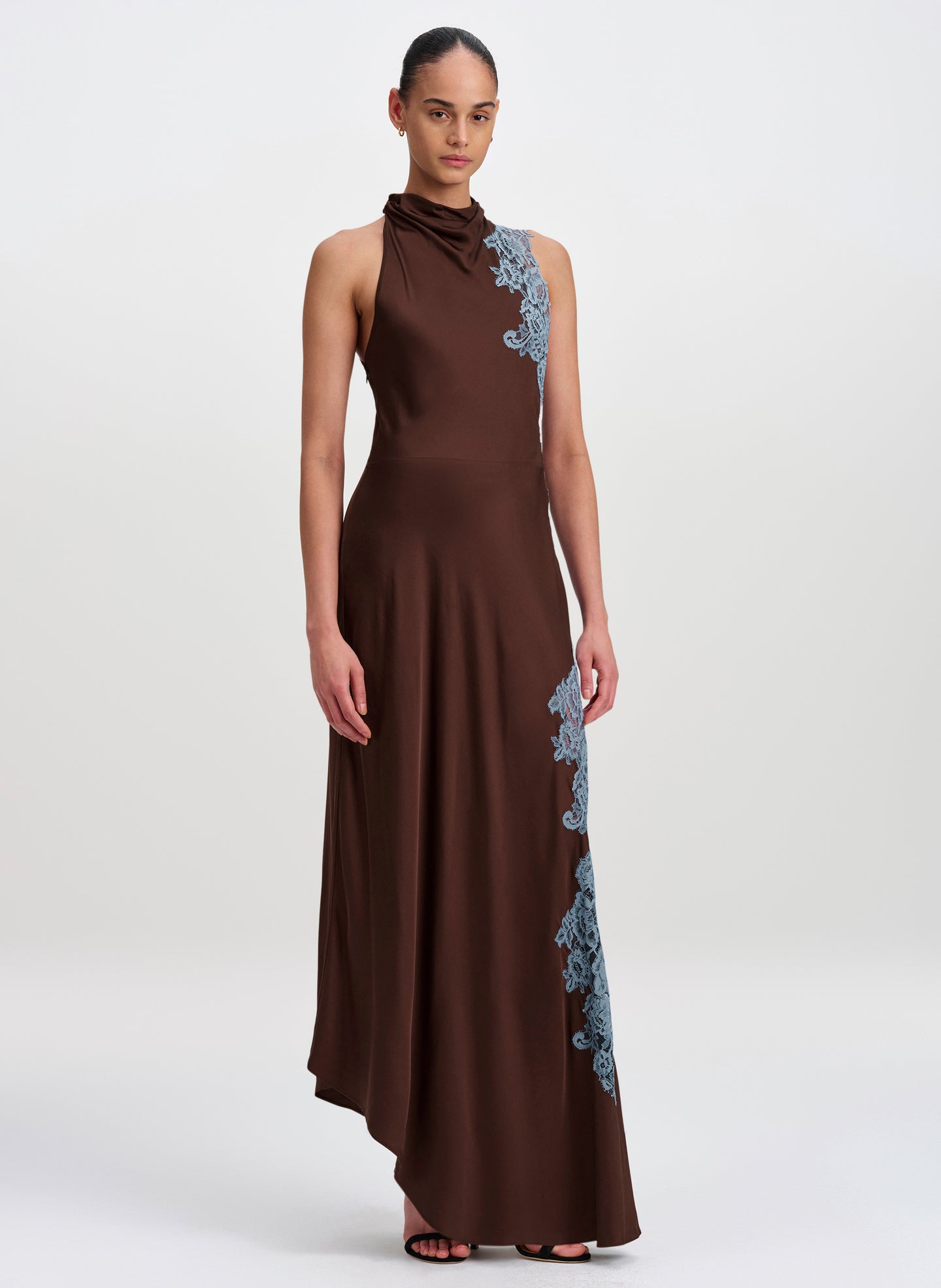 A model wearing a sleeveless, high-neck brown maxi dress with light blue floral lace applique detailing along the shoulder and side, set against a plain light gray studio background.