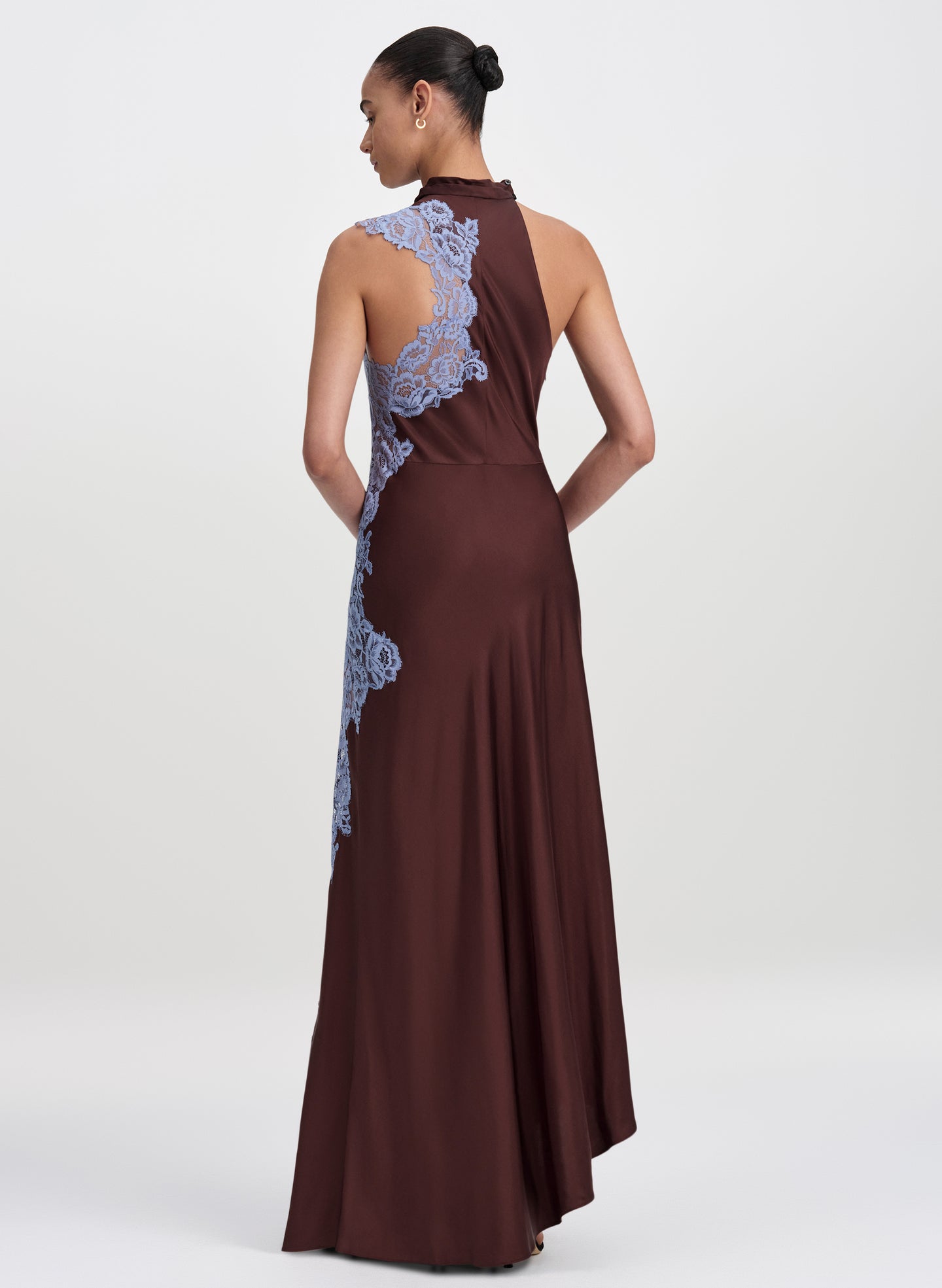 A woman modeling a floor-length sleeveless gown from the back, shown against a plain light gray studio background. The dress is dark brown with light blue floral lace applique running up the back and along the side.