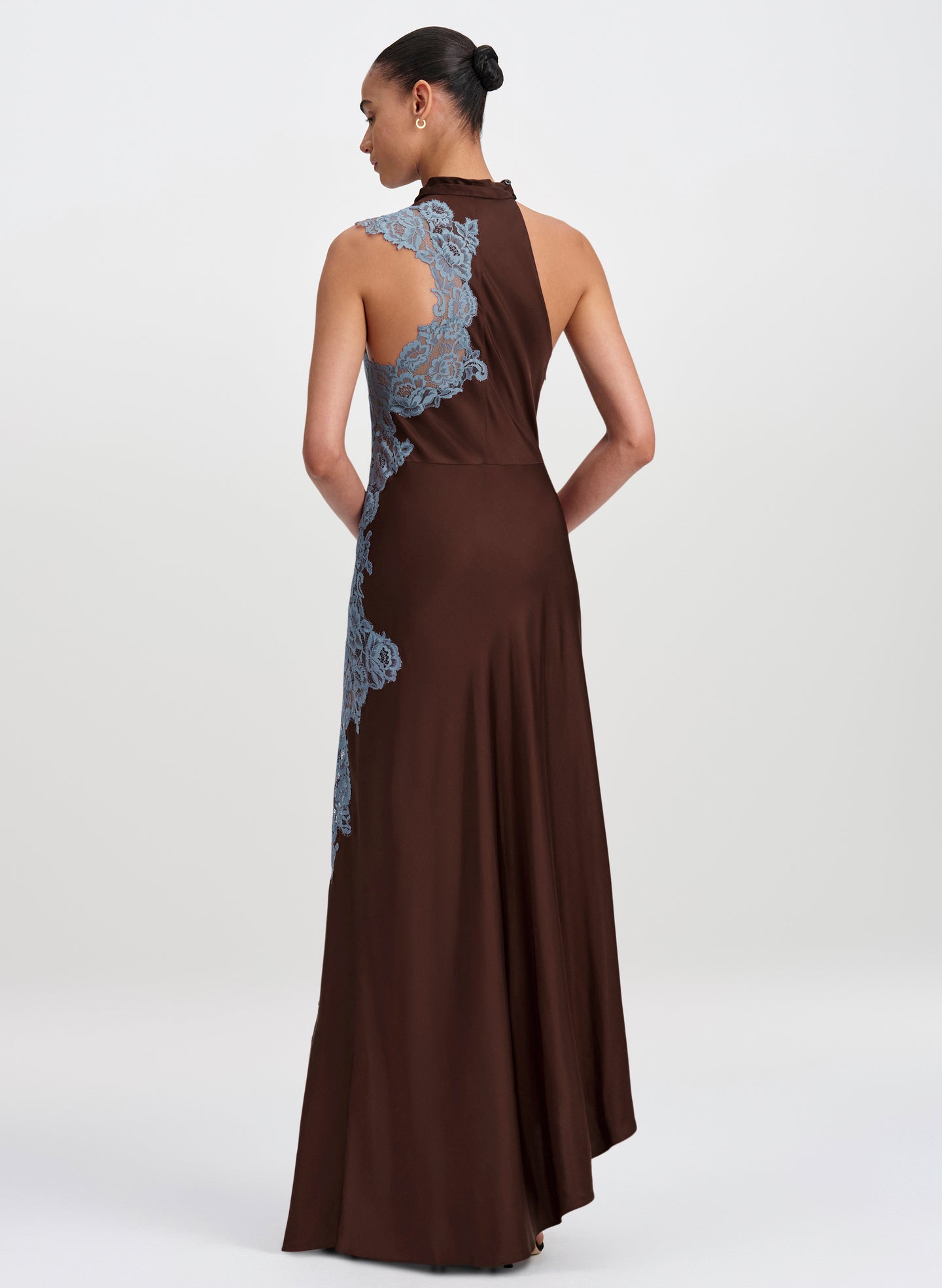 A woman modeling a floor-length brown dress from the back, featuring light blue lace applique panels along the back and side, set against a plain light gray studio background.