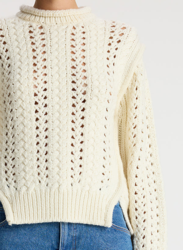 detail view of woman wearing cream open knit sweater and medium wash denim jeans