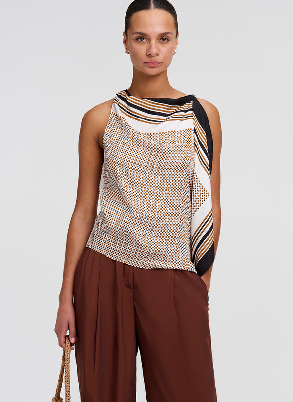 Woman wearing a sleeveless patterned top and brown pants on a plain background