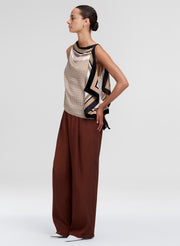 A model is shown in profile wearing a sleeveless top with a beige and black geometric pattern and wide-leg brown trousers, set against a plain light gray studio background.