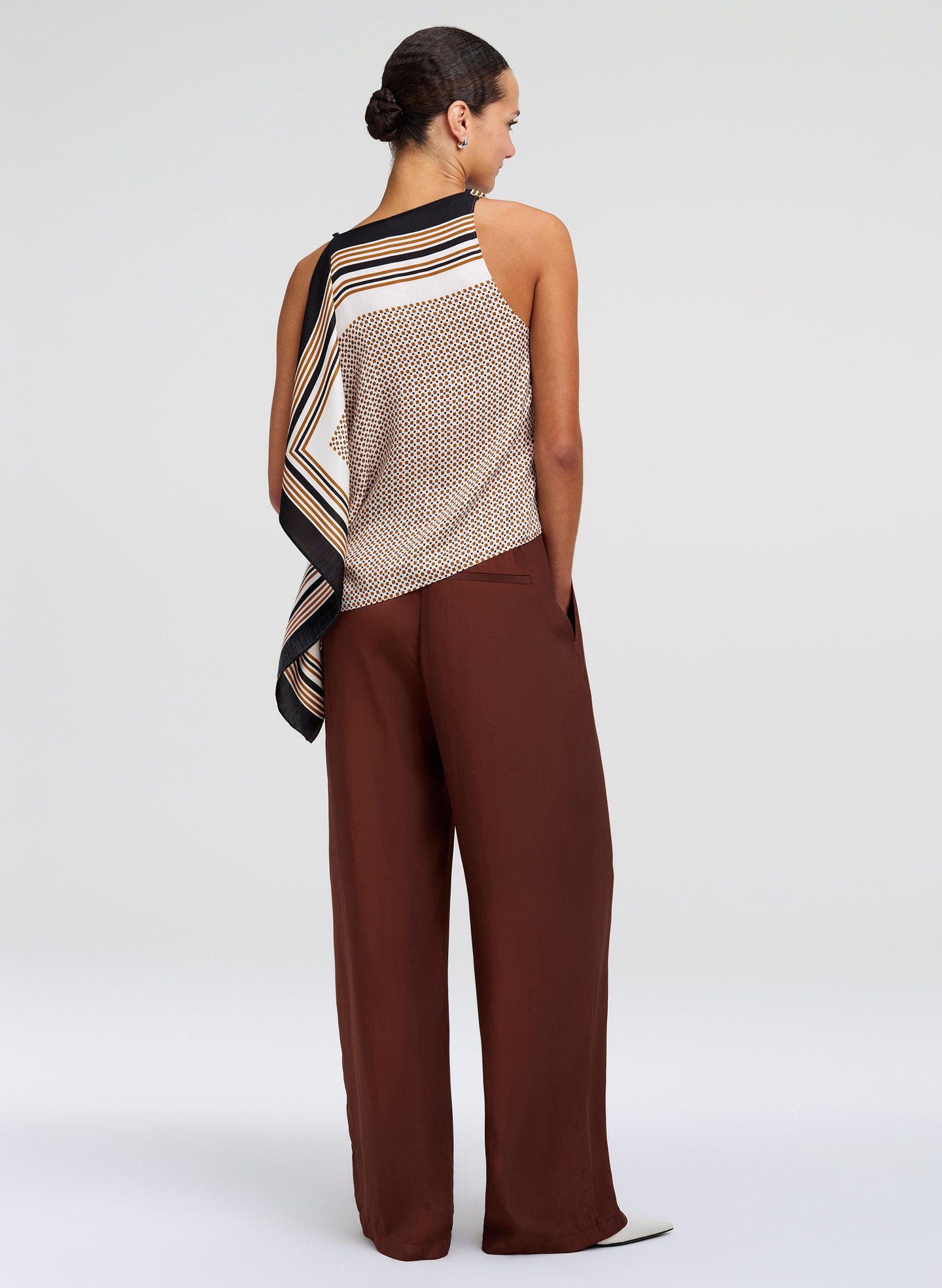A woman shown from the back wearing a sleeveless knit top with a striped scarf draped over one shoulder and wide-leg brown pants, photographed against a light gray studio background.