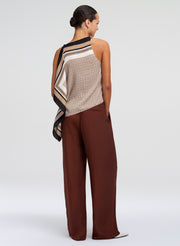 A woman shown from the back wearing a sleeveless knit top with a striped scarf draped over one shoulder and wide-leg brown pants, photographed against a light gray studio background.