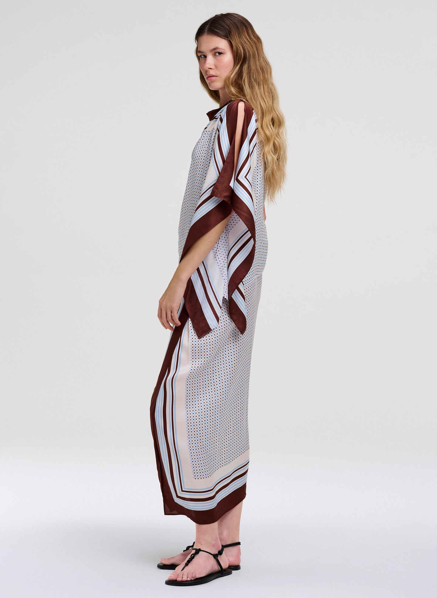 A woman modeling a long, loose midi skirt with matching top with a white base, brown and black striped geometric patterns, and a high collar, standing against a light gray studio background.