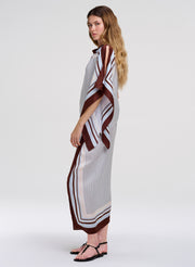 A woman modeling a long, loose midi skirt with matching top with a white base, brown and black striped geometric patterns, and a high collar, standing against a light gray studio background.