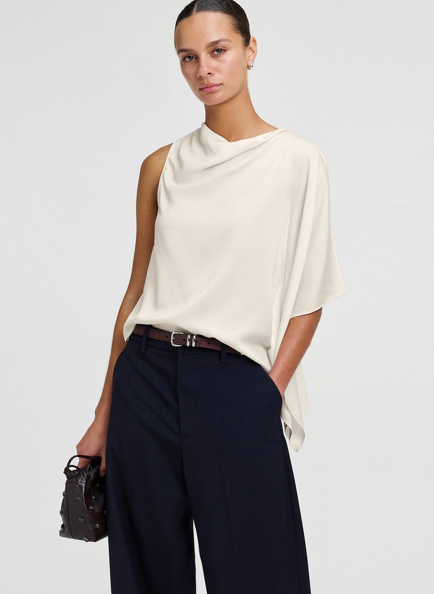A woman modeling a draped, sleeveless cream top paired with dark navy wide-leg trousers and a small black handbag, set against a plain light gray studio background.