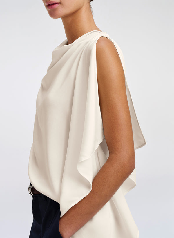 A close-up side view of a model wearing a sleeveless draped top in a warm off-white or cream color, paired with dark navy trousers, against a light gray studio background.