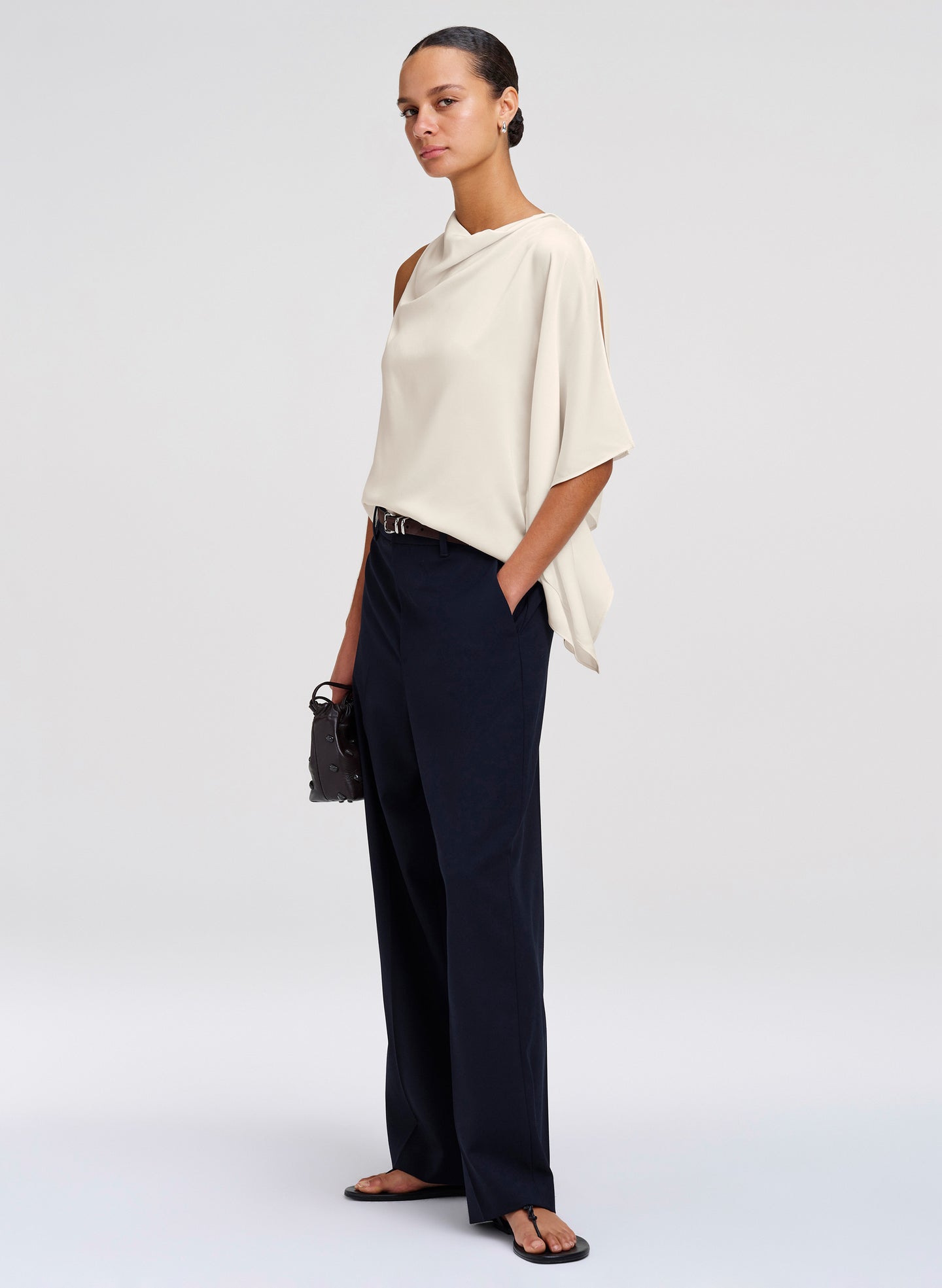 A model stands in a studio wearing a draped off-white top with cutout details, dark navy wide-leg trousers, black sandals, and holding a small black handbag.