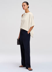 A model stands in a studio wearing a draped off-white top with cutout details, dark navy wide-leg trousers, black sandals, and holding a small black handbag.