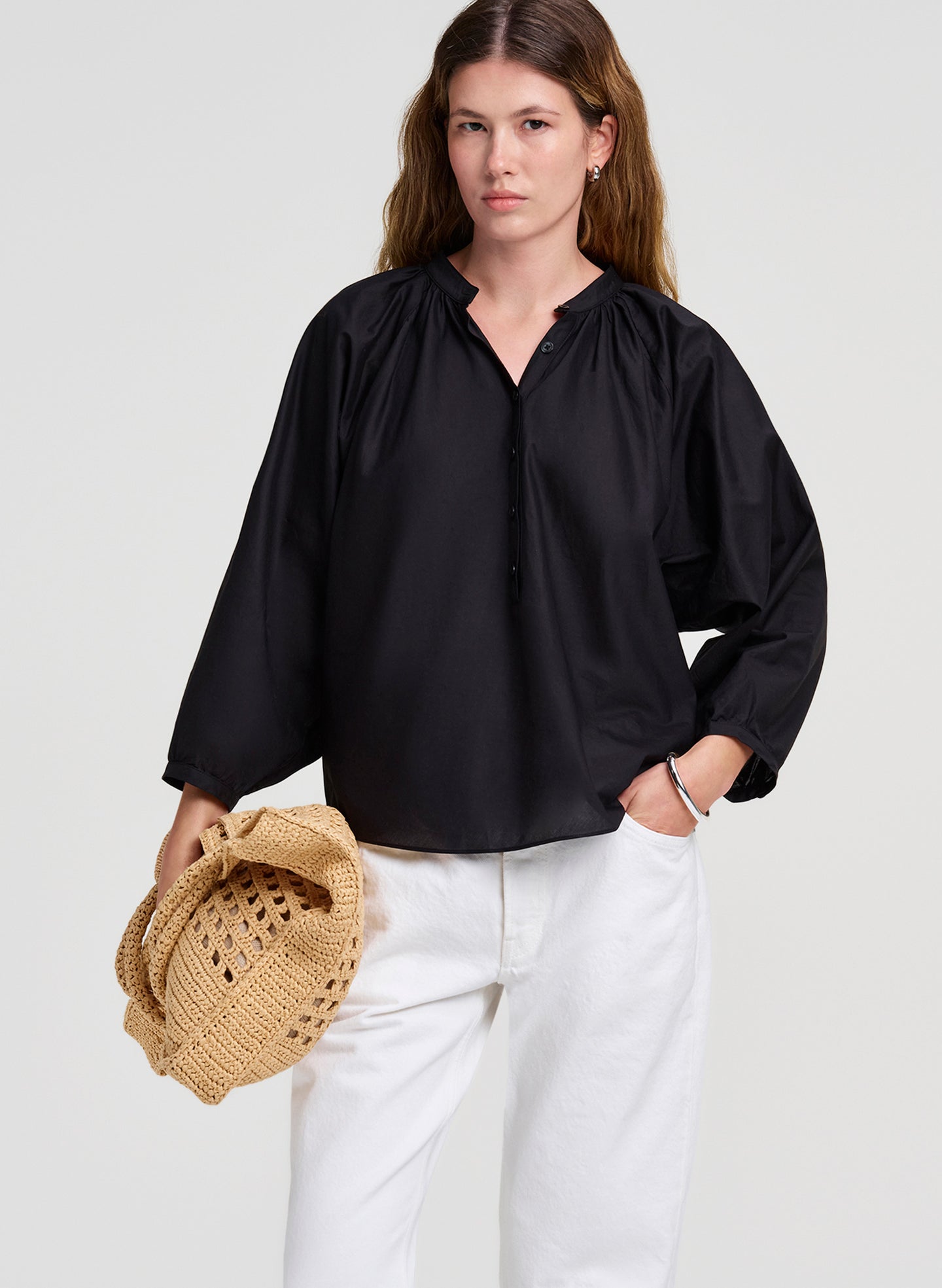 Woman wearing a black blouse and white pants holding a raffia handbag against a light gray background