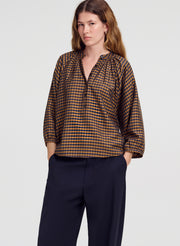 A woman modeling a brown and blue checkered long-sleeve blouse with a gathered neckline, paired with dark navy trousers, against a light gray studio background.