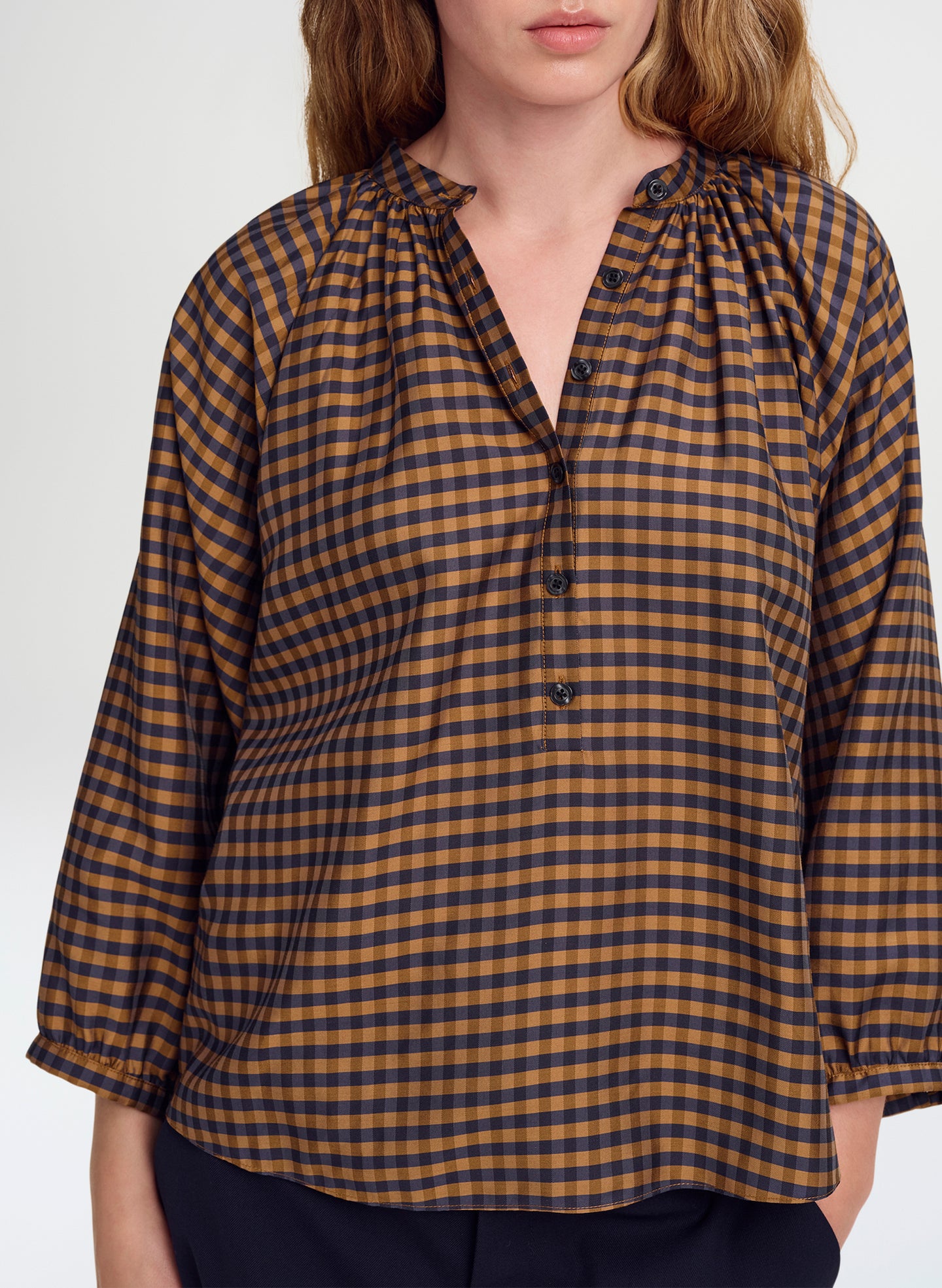 A woman wearing a brown and blue gingham check 3/4-sleeve blouse with a gathered, ruffled collar and button placket, paired with dark navy pants against a light gray studio background.