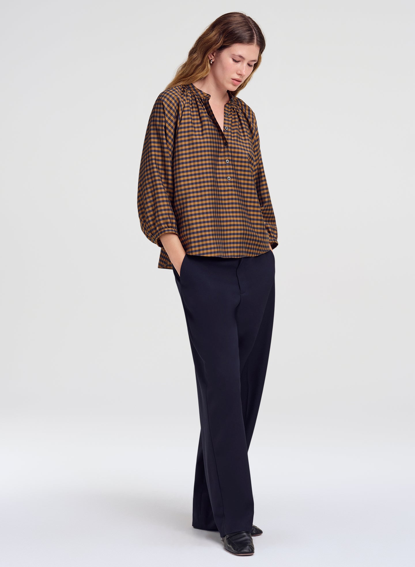 A woman modeling a brown and blue checkered long-sleeve blouse with dark navy wide-leg trousers, standing against a light gray studio background.