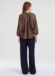 A woman shown from the back wearing a brown and blue checkered blouse with a gathered neckline and wide three-quarter sleeves, paired with dark wide-leg trousers, against a light gray studio background.
