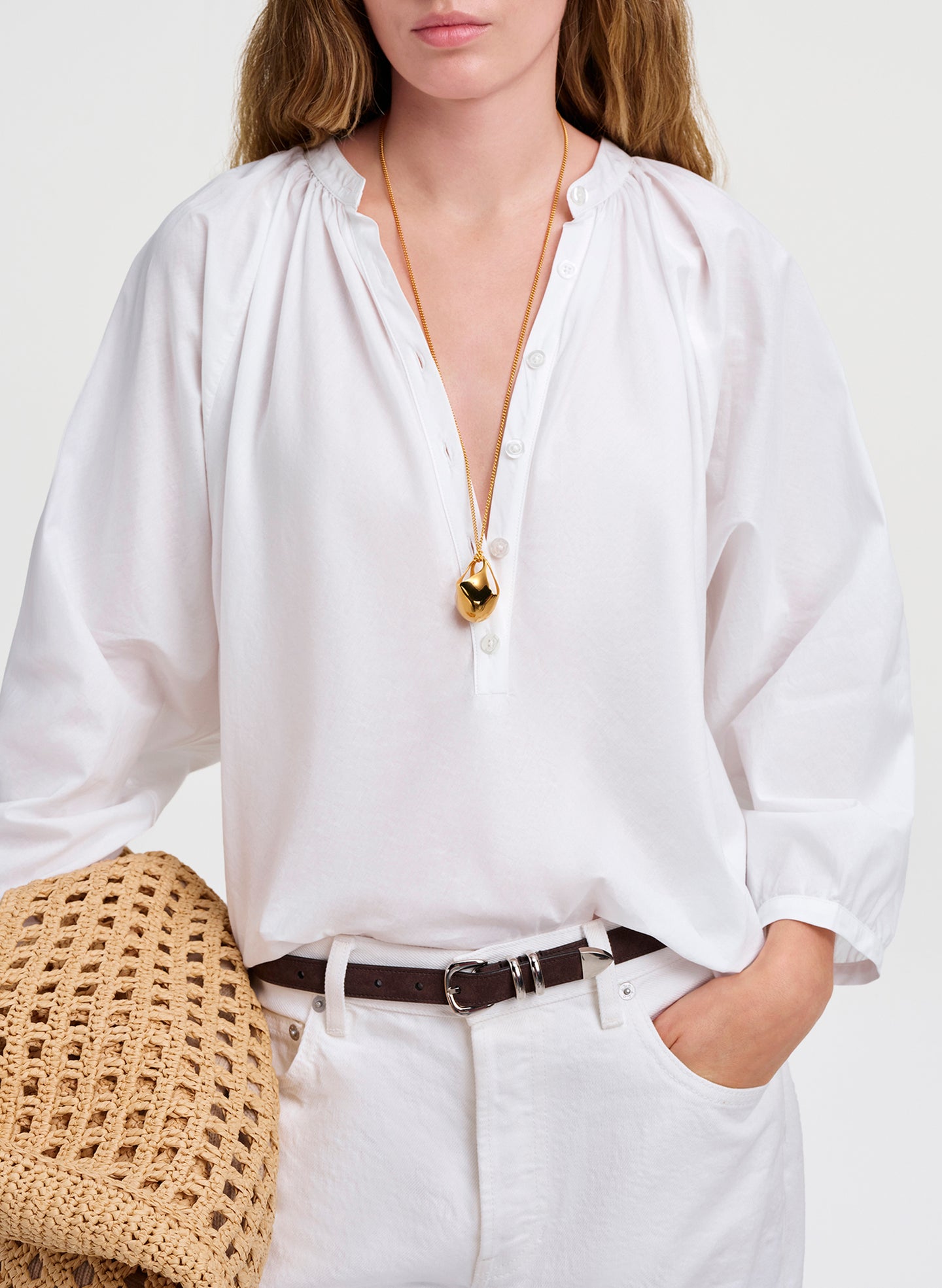 Person wearing a white shirt with a gold necklace and a woven bag on a light background
