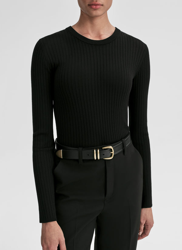 Person wearing a black ribbed long-sleeve top and black pants with a gold belt on a white background