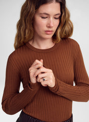 A woman wearing a ribbed long-sleeve crewneck sweater in a warm brown color, shown from the chest up against a light gray studio background.