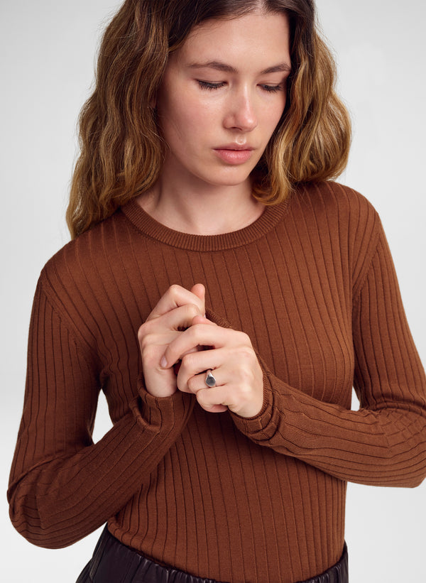 A woman wearing a ribbed long-sleeve crewneck sweater in a warm brown color, shown from the chest up against a light gray studio background.