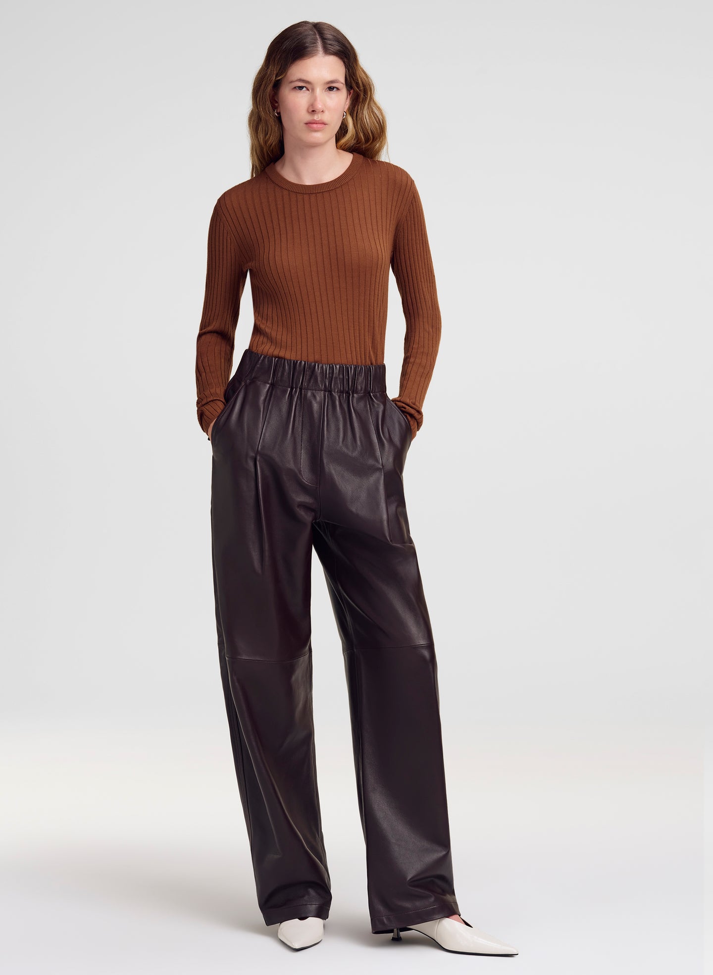 A model stands against a light gray studio background wearing a brown ribbed long-sleeve top and dark wide-leg pants with white pointed shoes.