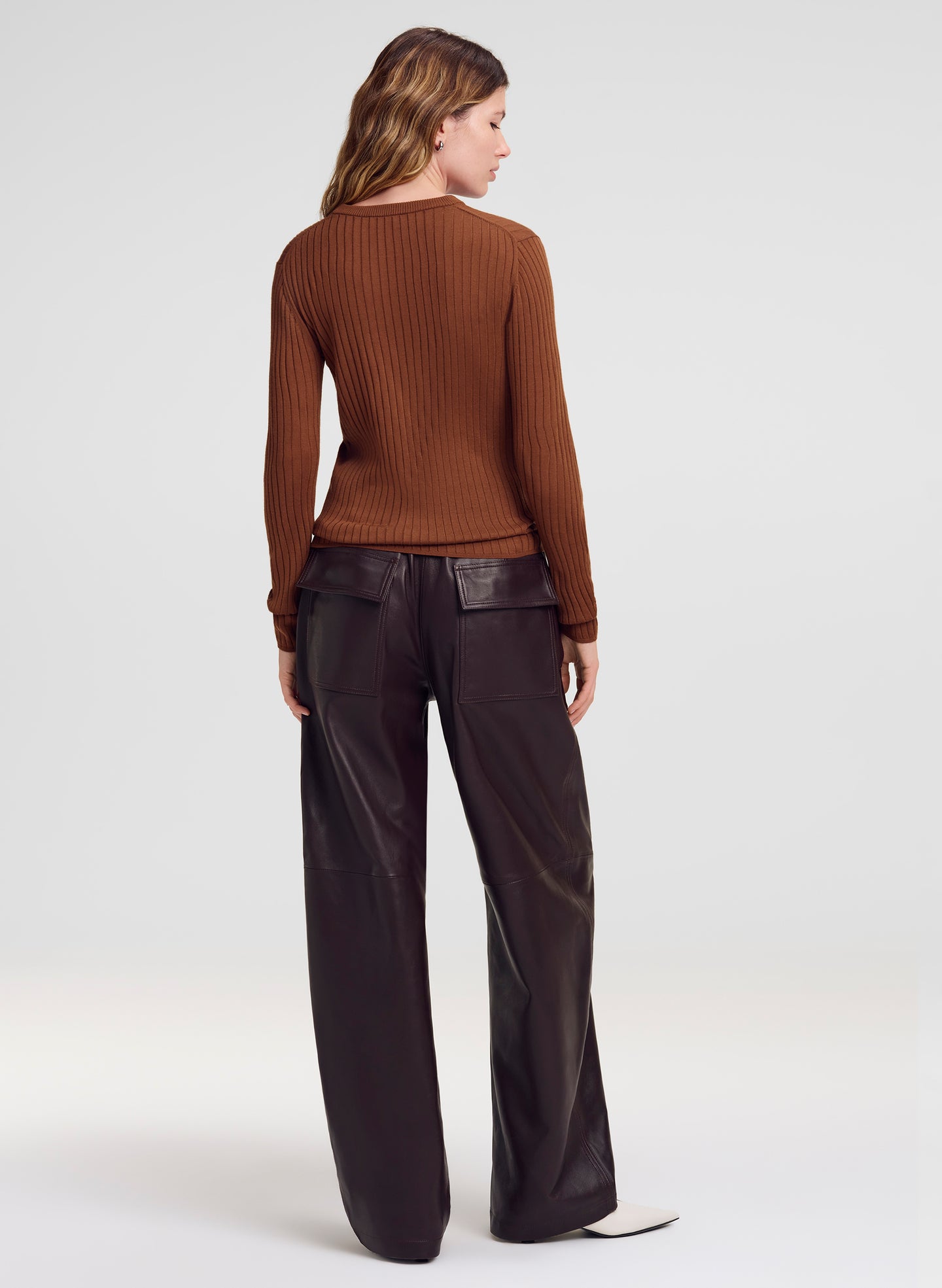 A woman shown from the back wearing a brown ribbed long-sleeve sweater and dark wide-leg pants against a light gray studio background.